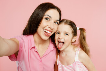 Wall Mural - Close up woman wear casual clothes with child kid girl 6-7 years old. Mother daughter do selfie shot pov on mobile cell phone show tongue isolated on plain pink background. Family parent day concept.