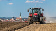 Red tractor in field at plowing stubble and harrowing dirt in Czech landscape. Farm vehicle working on arable land with view to church tower and historical center of Czech city Tabor under summer sky.