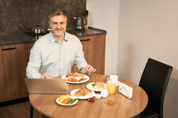 Portrait handsome man having breakfast with laptop in kitchen in hotel room