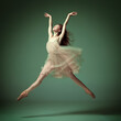 Young and incredibly beautiful ballerina wearing tulle dress jumping gracefully over dark green studio background. Demonstrate flexibility