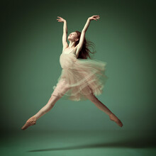 Young And Incredibly Beautiful Ballerina Wearing Tulle Dress Jumping Gracefully Over Dark Green Studio Background. Demonstrate Flexibility