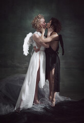 Portrait of two women, queens, angel and demon kissing against dark, green, vintage background. Sin love. Same-sex marriage. Concept of history, remake, good and bad, creative photography, lgbt