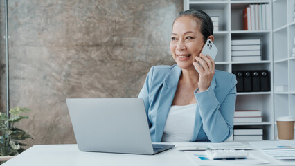 Confident asian chinese mature businesswoman working with laptop and desktop computer doing some paperwork at office workplace.