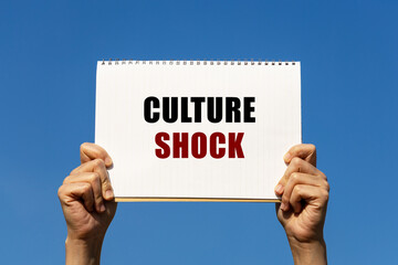 Culture shock text on notebook paper held by 2 hands with isolated blue sky background. This message can be used as business concept about culture shock.