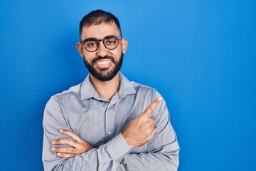 Poster - Middle east man with beard standing over blue background with a big smile on face, pointing with hand and finger to the side looking at the camera.