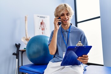 Wall Mural - Young blond man pysiotherapist reading document talking on smartphone at rehab clinic