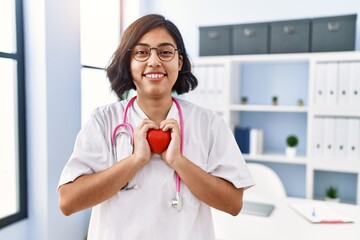 Canvas Print - Young latin woman wearing doctor uniform holding heart at clinic