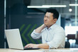 © Liubomir - A young man is an Asian worker wearing a white shirt works on a laptop in the office and holds his neck. Suffers and grimaces from pain, massages with his hand.