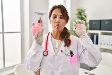 Hispanic Doctor Woman Holding Piggy Bank Making Fish Face With Mouth And Squinting Eyes, Crazy And Comical.