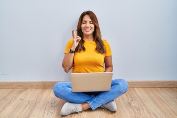 Sticker - Hispanic woman using laptop sitting on the floor at home gesturing finger crossed smiling with hope and eyes closed. luck and superstitious concept.