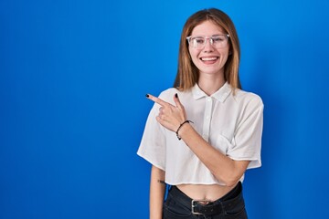 Poster - Beautiful woman standing over blue background cheerful with a smile of face pointing with hand and finger up to the side with happy and natural expression on face