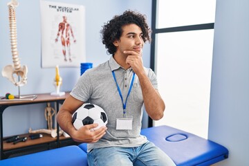 Poster - Hispanic man with curly hair working as football physiotherapist looking confident at the camera smiling with crossed arms and hand raised on chin. thinking positive.