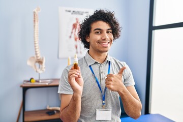 Canvas Print - Hispanic man with curly hair holding cbd oil smiling happy and positive, thumb up doing excellent and approval sign