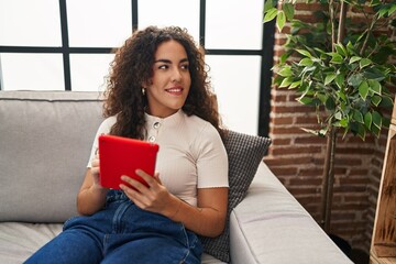 Wall Mural - Young beautiful hispanic woman using touchpad sitting on sofa at home