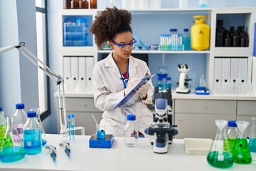 Canvas Print - Young african american woman wearing scientist uniform writing on clipboard at laboratory