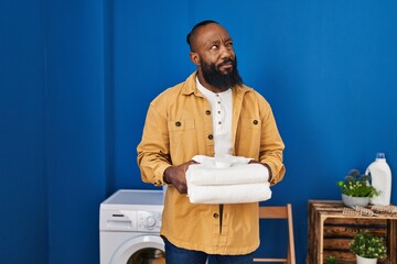 Poster - African american man holding clean towels at laundry room smiling looking to the side and staring away thinking.