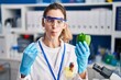 Young hispanic woman working at scientist laboratory with vegetables making fish face with mouth and squinting eyes, crazy and comical.