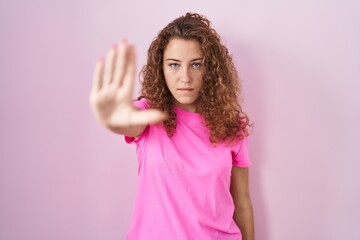 Wall Mural - Young caucasian woman standing over pink background doing stop sing with palm of the hand. warning expression with negative and serious gesture on the face.