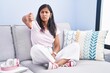 © Krakenimages.com - Young hispanic woman sitting on the sofa at home looking unhappy and angry showing rejection and negative with thumbs down gesture. bad expression.