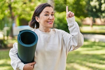 Middle age hispanic woman holding yoga mat at the park surprised with an idea or question pointing finger with happy face, number one