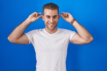 Canvas Print - Young caucasian man standing over blue background smiling pulling ears with fingers, funny gesture. audition problem