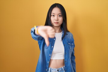 Canvas Print - Young asian woman standing over yellow background looking unhappy and angry showing rejection and negative with thumbs down gesture. bad expression.