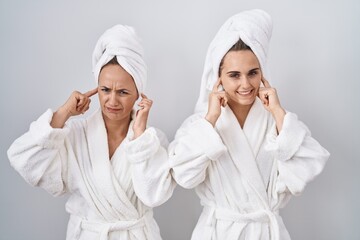 Canvas Print - Middle age woman and daughter wearing white bathrobe and towel covering ears with fingers with annoyed expression for the noise of loud music. deaf concept.