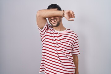 Wall Mural - Hispanic man with long hair standing over isolated background covering eyes with arm smiling cheerful and funny. blind concept.