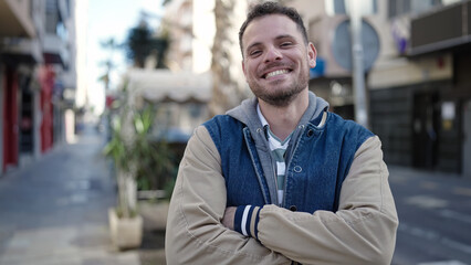 Wall Mural - Young caucasian man smiling with crossed arms at street