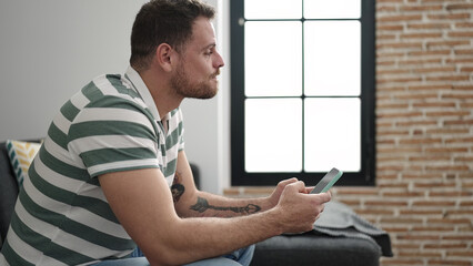 Canvas Print - Young caucasian man using the phone with serious expression sitting on the sofa at home