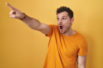 Wall Mural - Young hispanic man standing over yellow background pointing with finger surprised ahead, open mouth amazed expression, something on the front