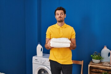 Poster - Young hispanic man holding clean towels at laundry room smiling looking to the side and staring away thinking.