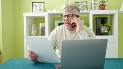 Wall Mural - Middle age grey-haired man talking on smartphone reading document at home