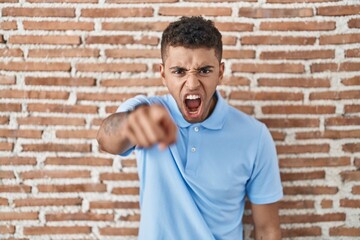 Sticker - Brazilian young man standing over brick wall pointing displeased and frustrated to the camera, angry and furious with you