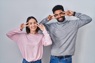 Poster - Young hispanic couple standing together doing peace symbol with fingers over face, smiling cheerful showing victory