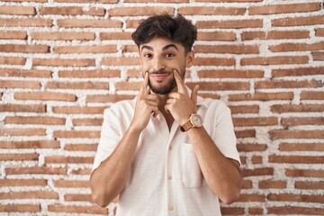 Sticker - Arab man with beard standing over bricks wall background smiling with open mouth, fingers pointing and forcing cheerful smile