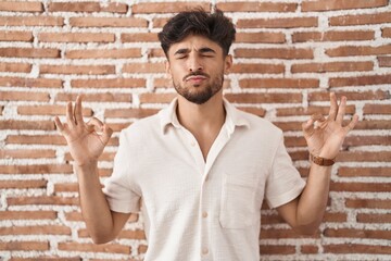 Canvas Print - Arab man with beard standing over bricks wall background relaxed and smiling with eyes closed doing meditation gesture with fingers. yoga concept.