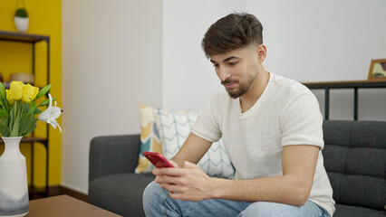 Wall Mural - Young arab man using smartphone sitting on sofa at home