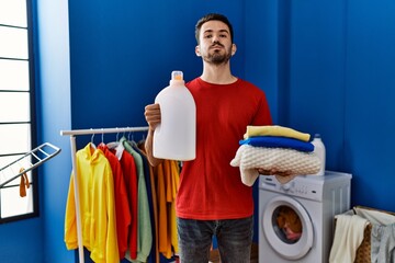 Canvas Print - Young hispanic man holding detergent bottle puffing cheeks with funny face. mouth inflated with air, catching air.