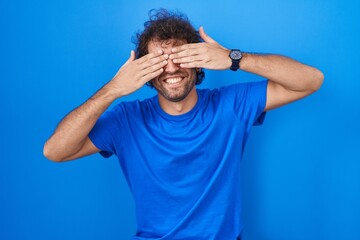 Canvas Print - Hispanic young man standing over blue background covering eyes with hands smiling cheerful and funny. blind concept.