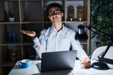 Wall Mural - Hispanic man working at the office at night smiling cheerful presenting and pointing with palm of hand looking at the camera.