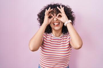 Sticker - Young middle east woman standing over pink background doing ok gesture like binoculars sticking tongue out, eyes looking through fingers. crazy expression.