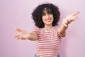 Sticker - Young middle east woman standing over pink background smiling cheerful offering hands giving assistance and acceptance.