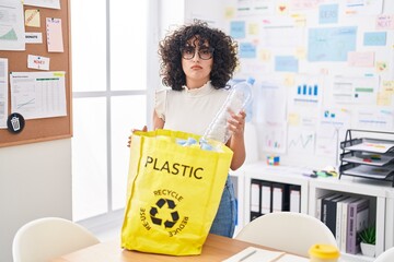 Poster - Young middle east woman holding recycling bag with plastic bottles at the office relaxed with serious expression on face. simple and natural looking at the camera.