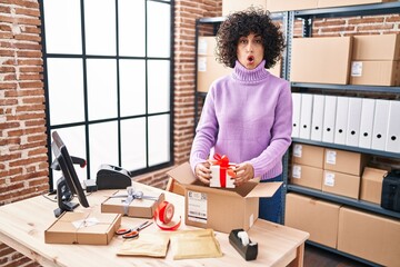 Sticker - Young brunette woman with curly hair working at small business ecommerce preparing order scared and amazed with open mouth for surprise, disbelief face