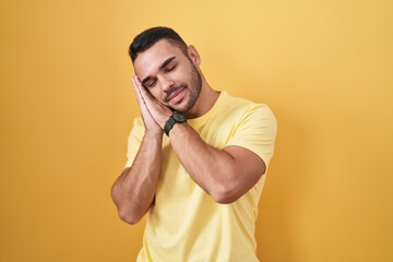 Canvas Print - Young hispanic man standing over yellow background sleeping tired dreaming and posing with hands together while smiling with closed eyes.