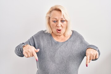 Middle age caucasian woman standing over white background pointing down with fingers showing advertisement, surprised face and open mouth