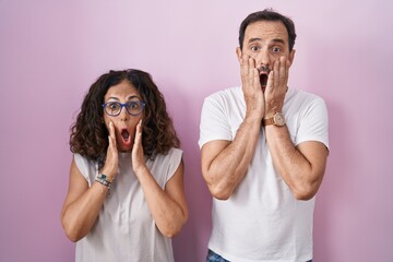 Poster - Middle age hispanic couple together over pink background afraid and shocked, surprise and amazed expression with hands on face