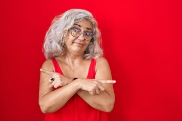 Sticker - Middle age woman with grey hair standing over red background pointing to both sides with fingers, different direction disagree