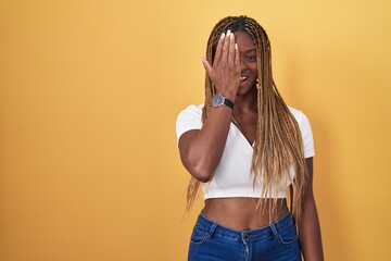 Wall Mural - African american woman with braided hair standing over yellow background covering one eye with hand, confident smile on face and surprise emotion.